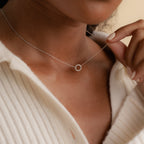 A woman wears the Pave Interlocking Necklace—a delicate silver jewelry piece with a small circular pendant—holding it gently near her collar.