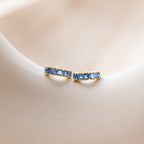 A pair of gold Genevieve Birthstone Huggies featuring square-cut lblue topaz gemstones, elegantly set in a row to form a sleek hoop design, displayed on a soft neutral background.