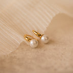 A close-up product shot of a pair of elegant gold-tone huggie earrings, each featuring a smooth, round white pearl drop.