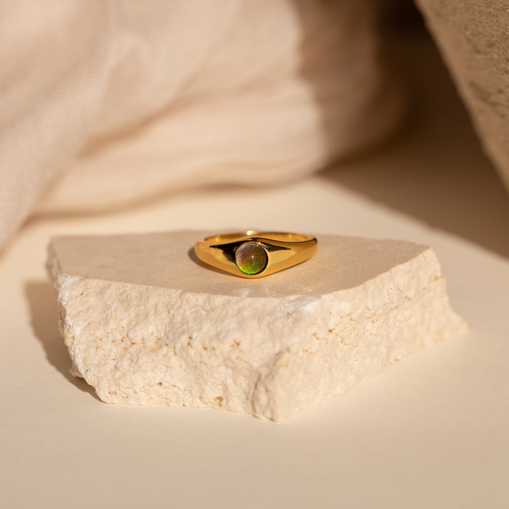 The Signet Mood Ring features a color-changing green gemstone on a textured white stone, set against a soft beige background—nostalgic jewelry with a modern twist.
