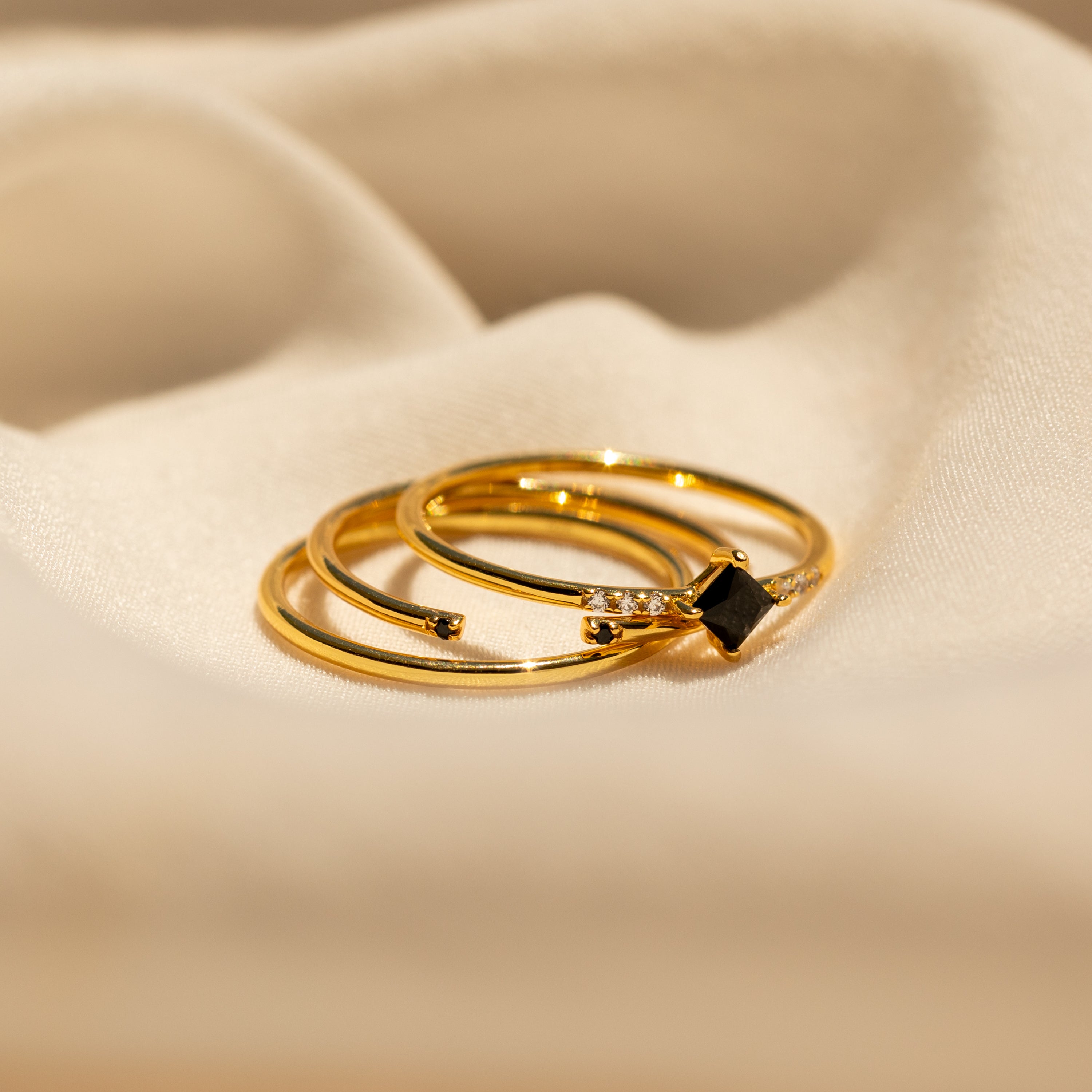 The Black Abyss Ring Set features three gold dainty bands with black gemstones and small diamonds, displayed on a soft beige fabric background.