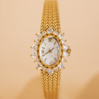 The Pave Heart Timepiece features a diamond-trimmed oval face and a woven gold band, elegantly displayed on a beige background.