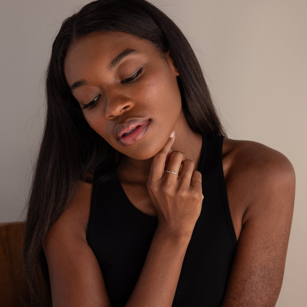 A woman with long dark hair, wearing a black tank top and looking down, touches her chin while displaying the Alex Dainty Letter Ring in Sterling Silver—a personalized piece that adds subtle elegance to her look.