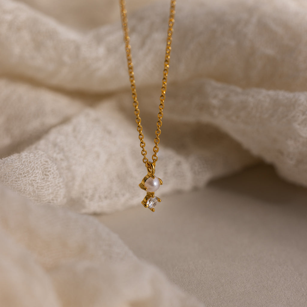 The Pearl Diamond Drop Necklace features a petite pearl and diamond pendant—an elegant bridal jewelry piece displayed on soft, cream-colored fabric.