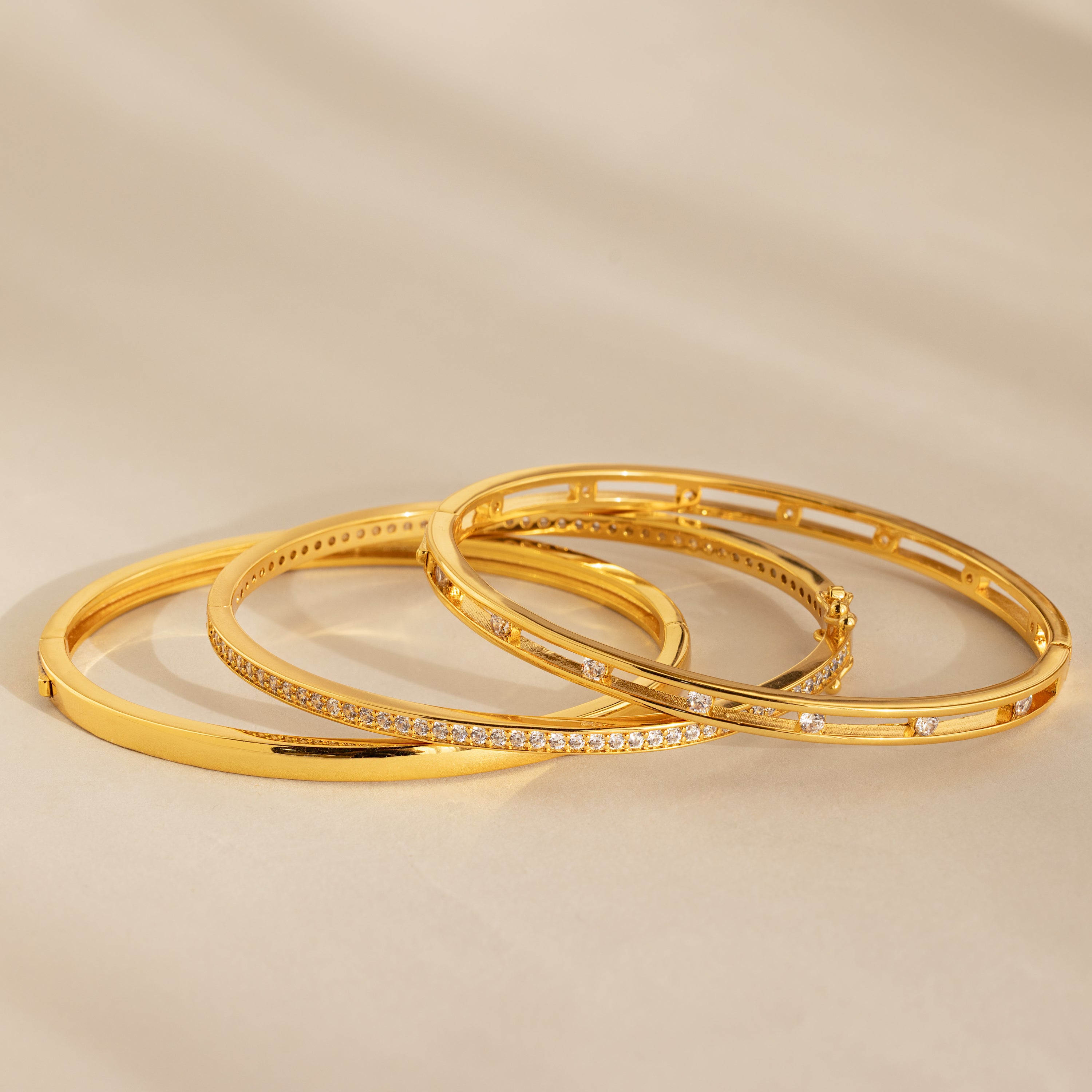Three gold bangles, including the Lace Diamond Bangle in 18K Gold with round diamonds and an open station design, are displayed on a neutral beige surface.