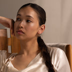Woman with braided hair in a satin top sits on a wooden chair, gazing thoughtfully to the side while her Inside Out Pear Diamond Hoops in 18K Gold add an elegant touch to her look.