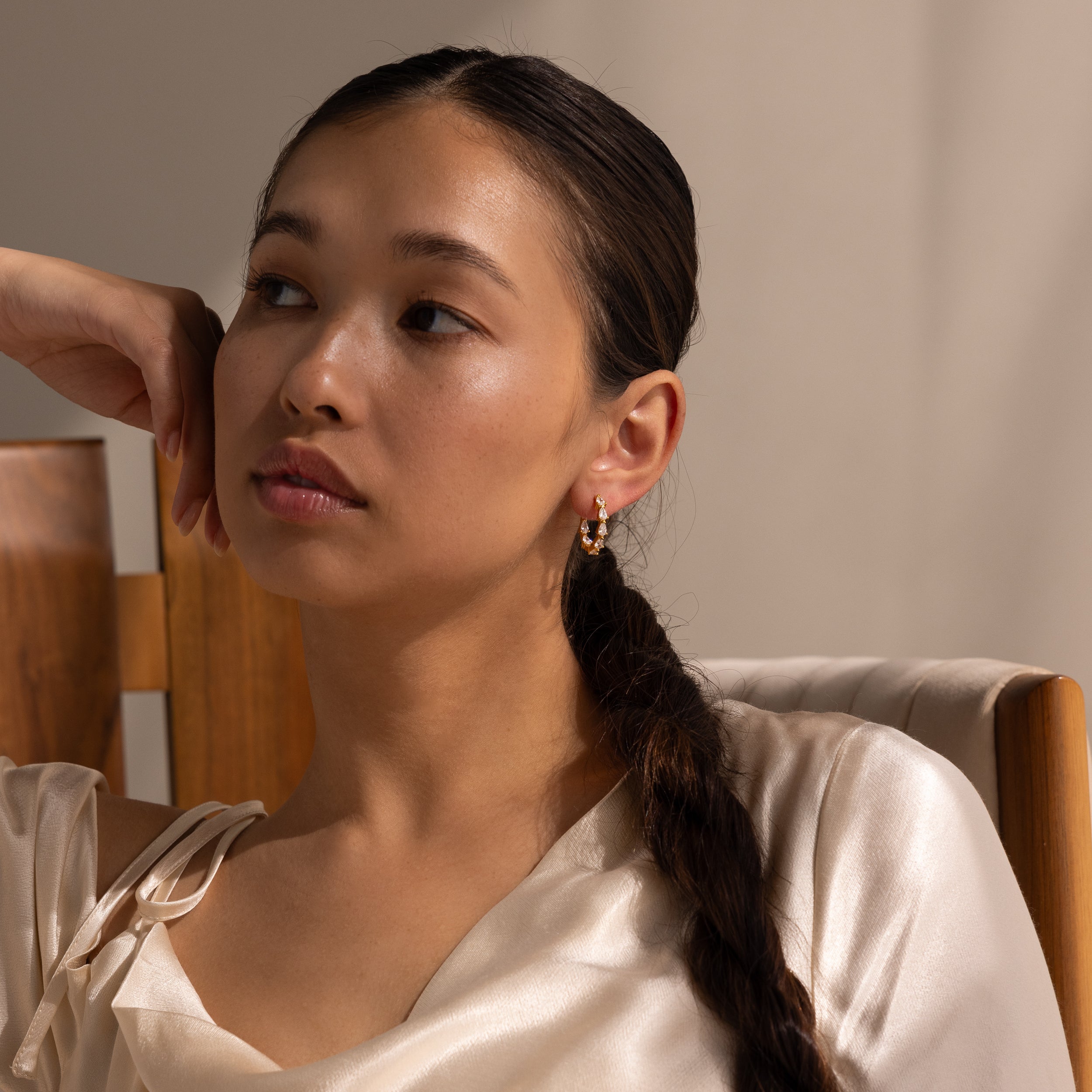 Woman with braided hair in a satin top sits on a wooden chair, gazing thoughtfully to the side while her Inside Out Pear Diamond Hoops in 18K Gold add an elegant touch to her look.