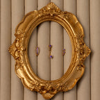 The Amethyst Earring Trio Set features three small purple birthstone earrings in a gold ornate oval frame on a beige padded background—an elegant gold set, perfect for gifting.