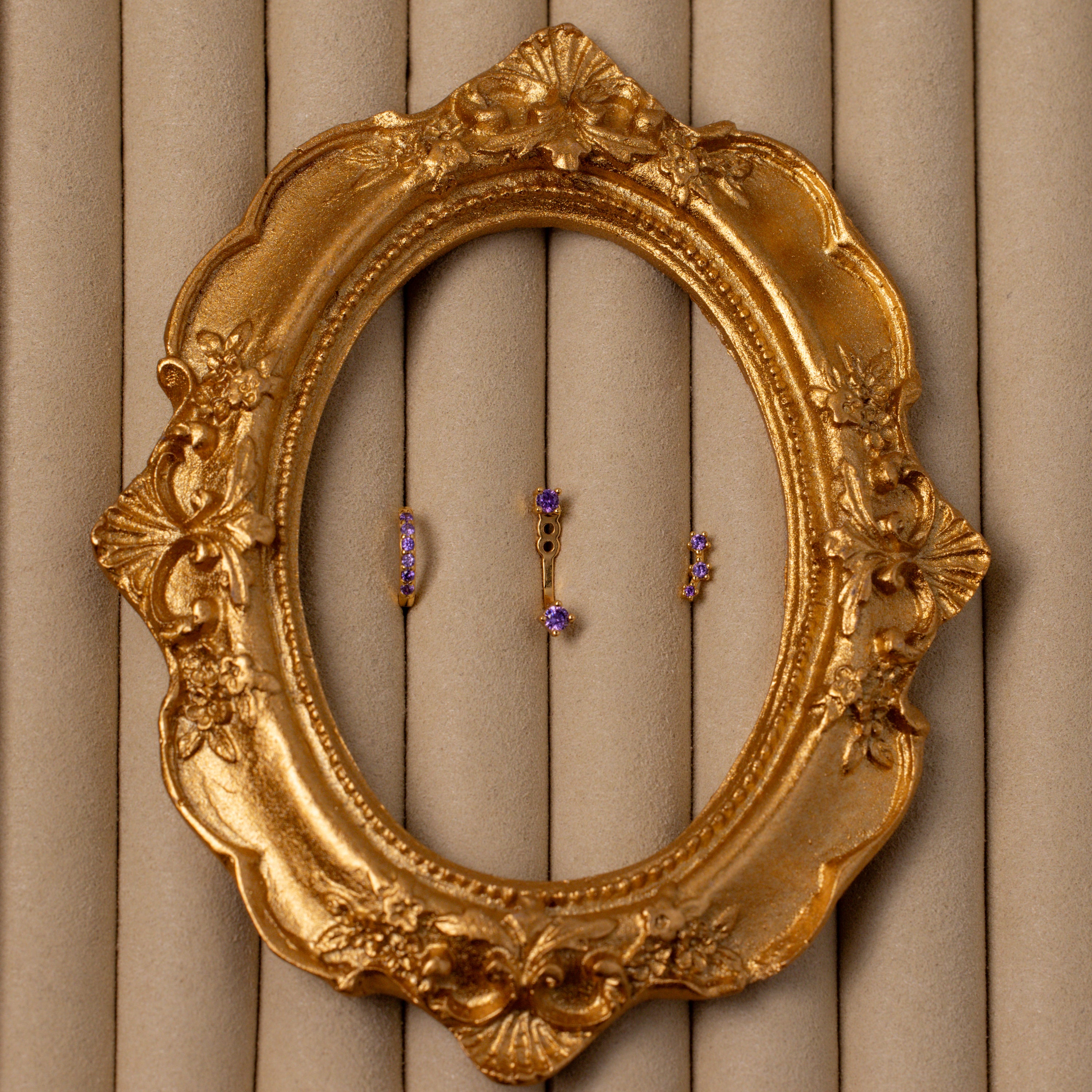 The Birthstone Earring Trio Set, featuring three small gold and purple earrings, is displayed inside an ornate oval gold frame on a beige textured background.
