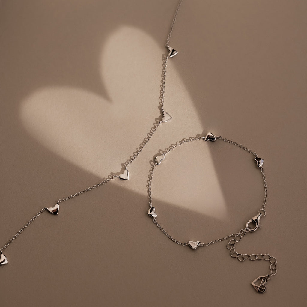 The Heart Station Necklace in Silver features delicate heart charms and rests on a beige surface, casting romantic, heart-shaped shadows in the background.
