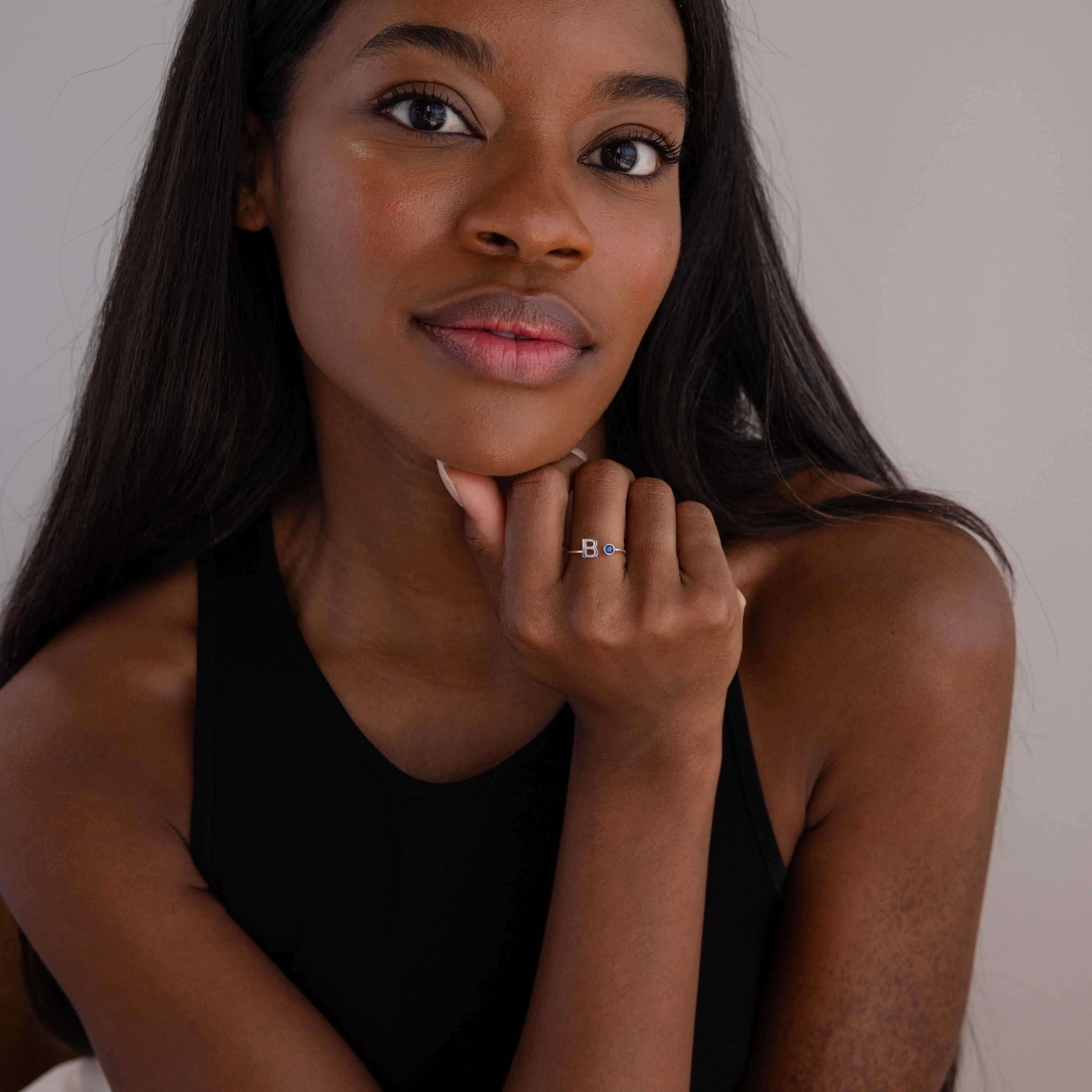 A woman with long dark hair in a black top rests her chin on her hand, showcasing the Alex Initial Birthstone Ring in Sterling Silver.