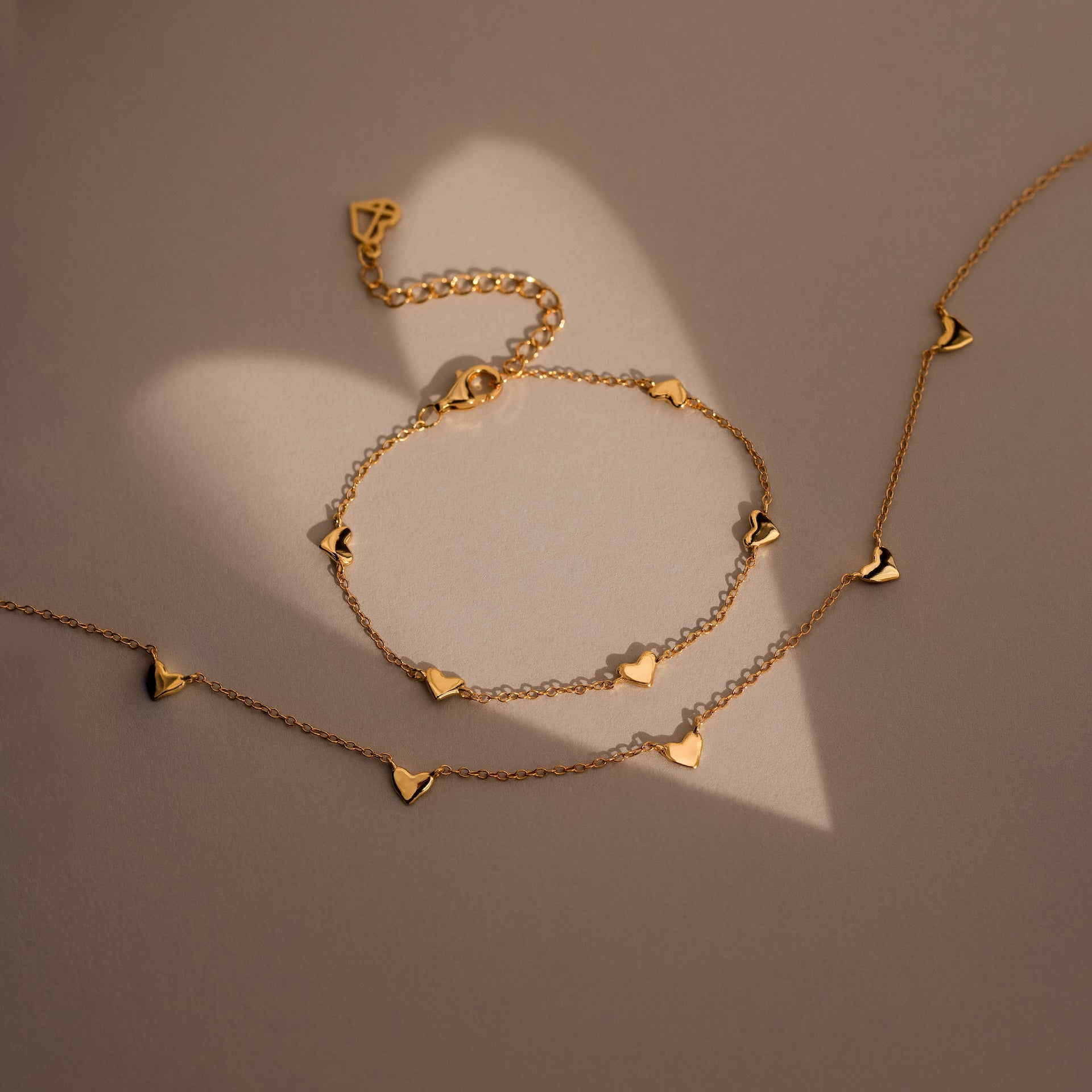Heart Station Necklace with delicate heart charms on a cable chain, displayed on a beige surface casting a heart-shaped shadow.