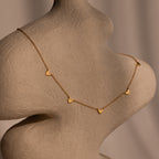 The Heart Station Necklace features delicate heart charms on a classic cable chain, showcased on a textured beige mannequin bust under soft lighting.