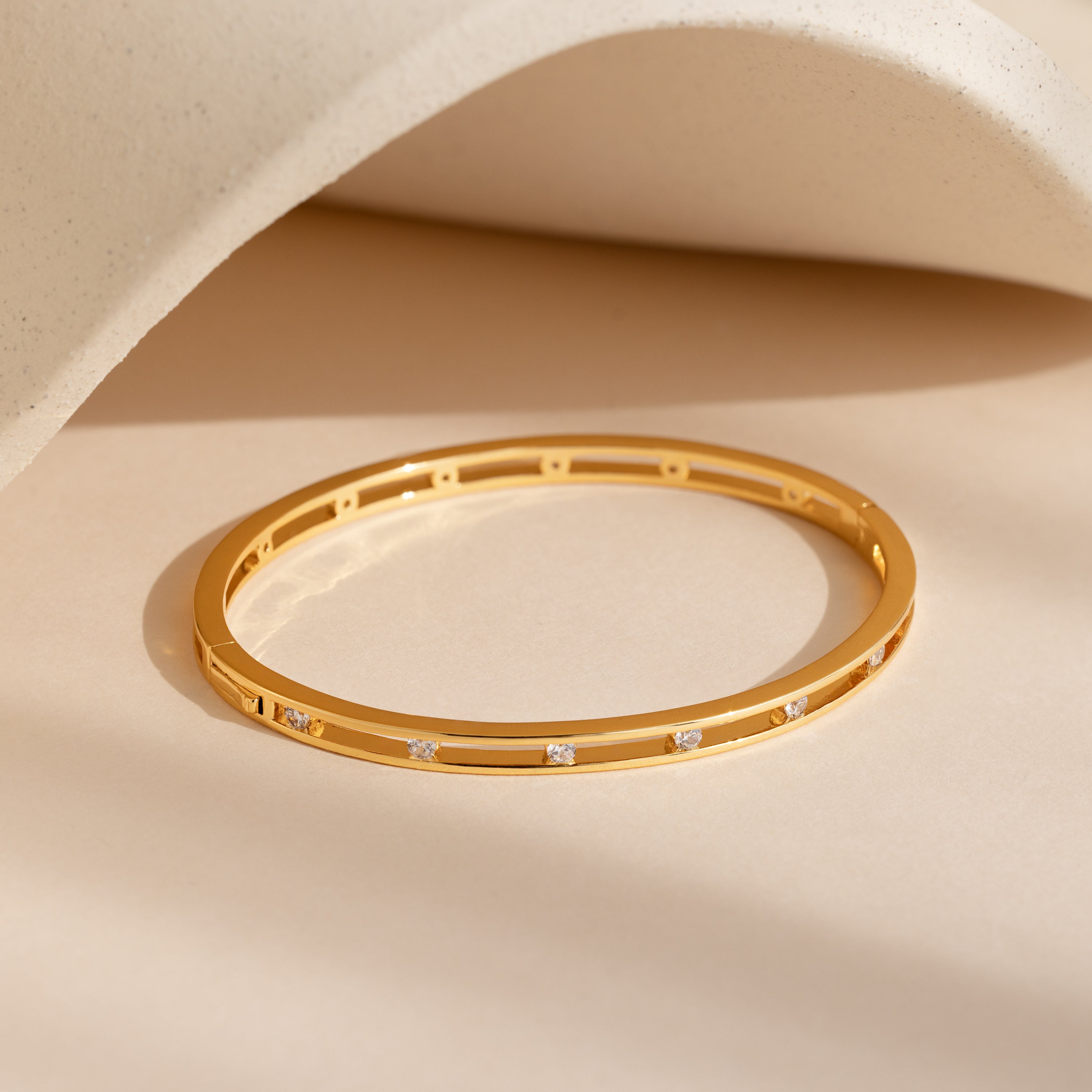 The Lace Diamond Bangle in 18K Gold has an open station design adorned with small round diamonds, elegantly displayed on a beige surface with a curved background.