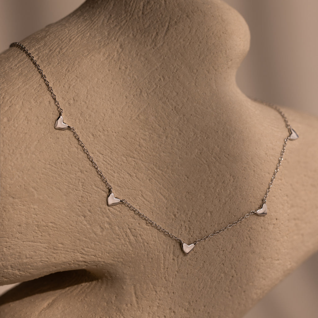 Heart Station Necklace in Silver, featuring delicate heart charms on a cable chain, displayed on a beige textured mannequin bust.