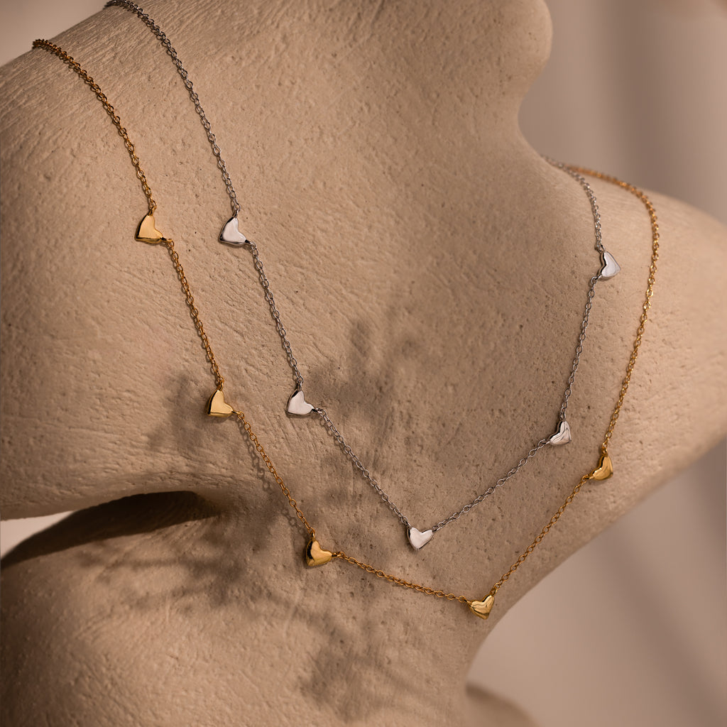 Two Heart Station Necklaces—one gold, one silver—each with a small heart charm, are displayed on a beige mannequin, both suspended from sleek cable chains.