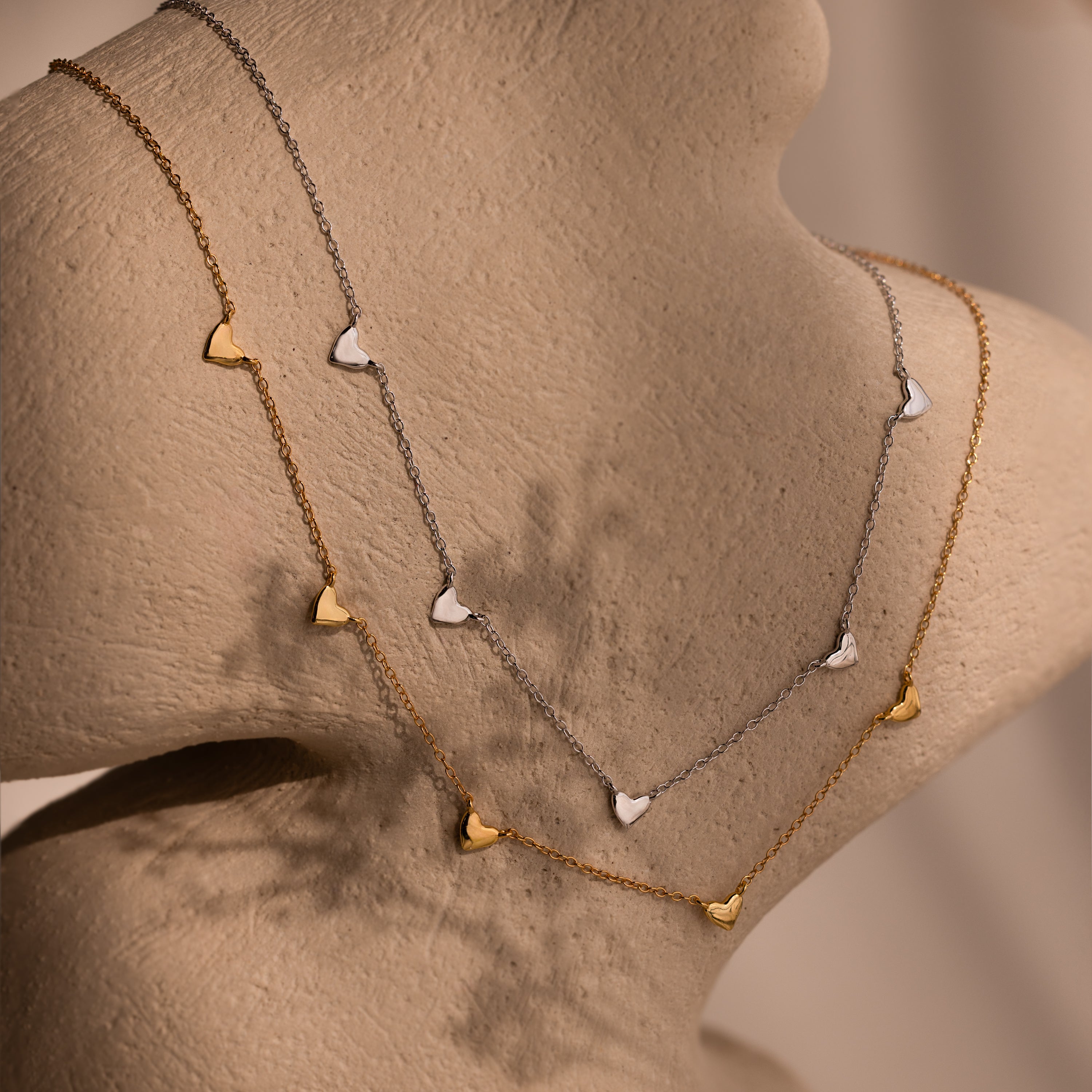 Two Heart Station Necklaces—one gold, one silver—each with a small heart charm, are displayed on a beige mannequin, both suspended from sleek cable chains.