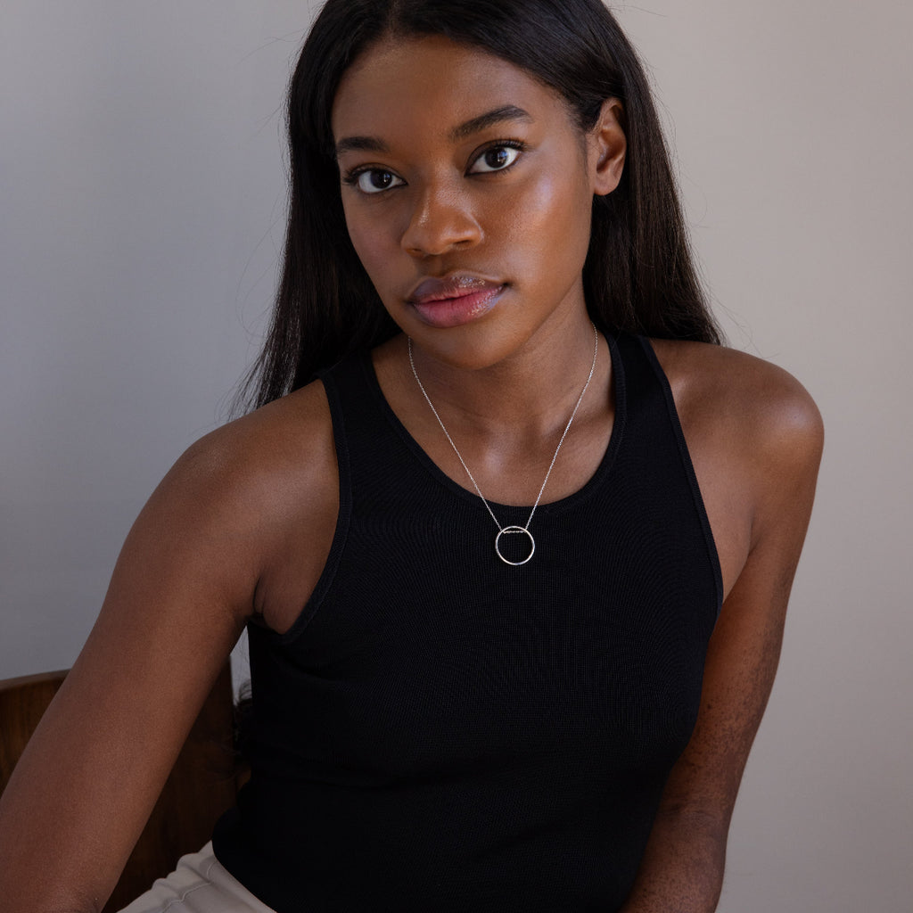 A woman with long dark hair, wearing a black sleeveless top and the Inner Circle Necklace in Sterling Silver—a striking silver circle pendant and chain—sits indoors.