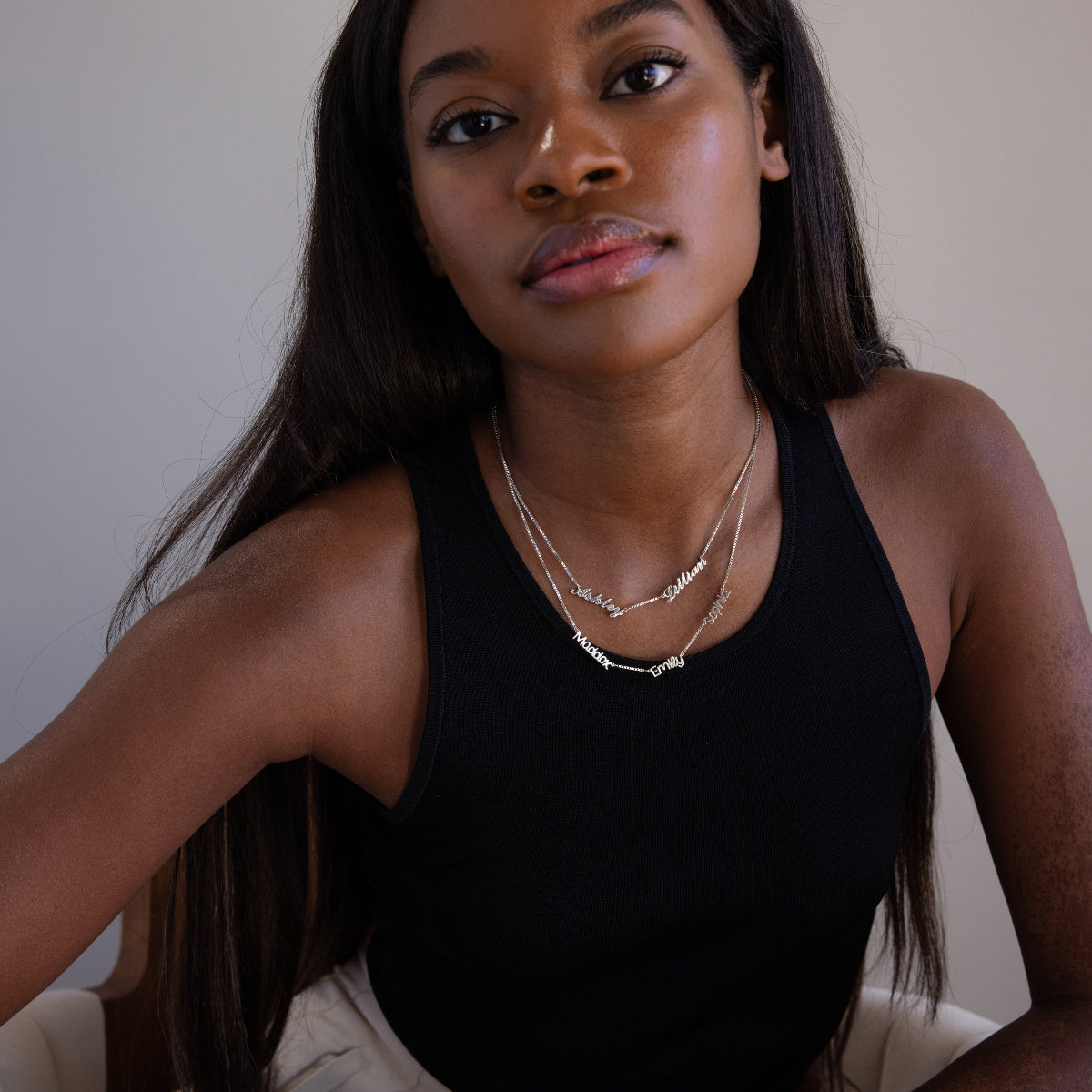 Woman wearing a black tank top and layered silver multiple name necklaces against a neutral background