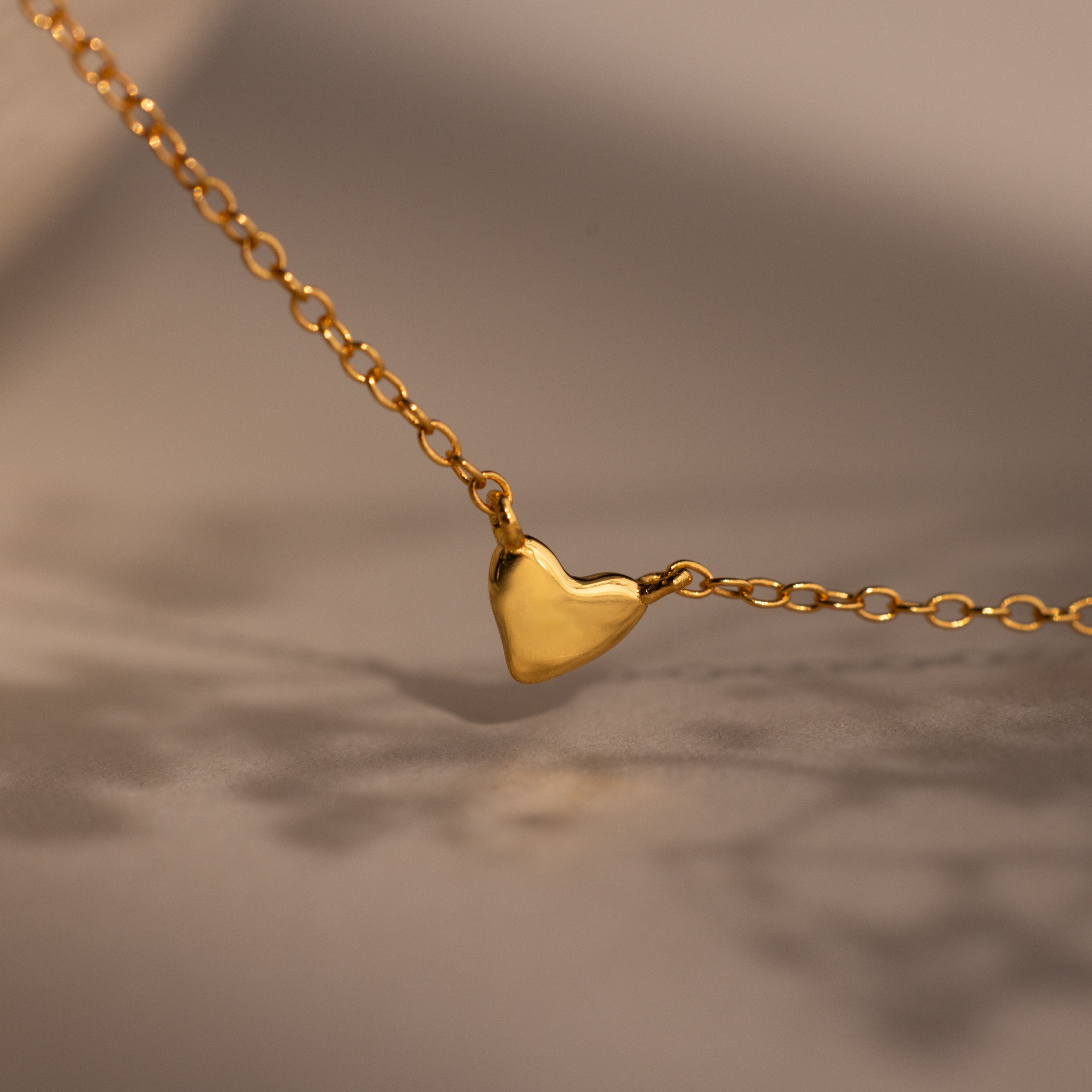 The Heart Station Necklace features a small, smooth gold heart pendant displayed on a soft beige background.