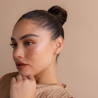 A woman with dark hair in a bun, wearing Opal Flat Back Studs, rests her chin on her hand and gazes to the side against a beige background.