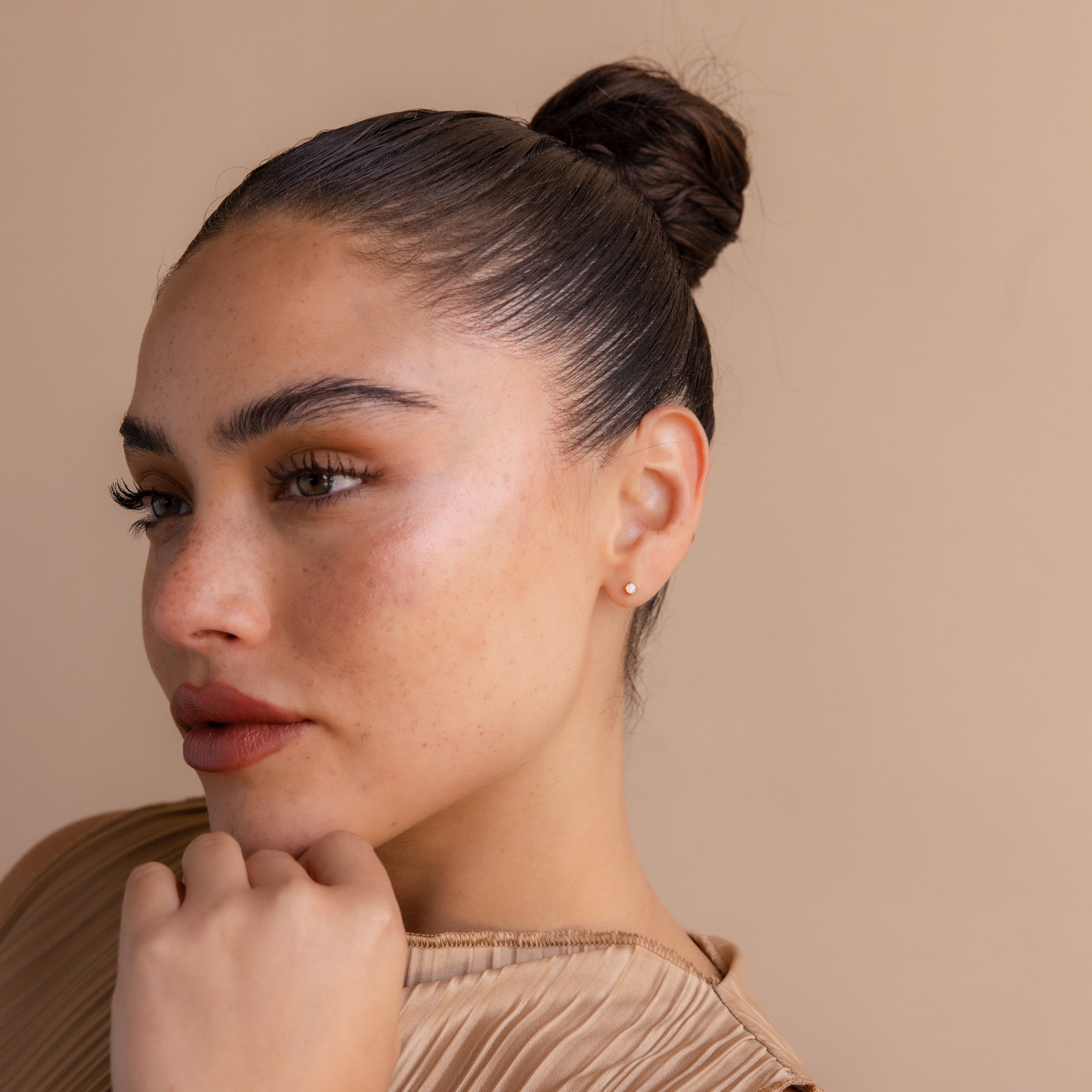 A woman with dark hair in a bun, wearing Opal Flat Back Studs, rests her chin on her hand and gazes to the side against a beige background.
