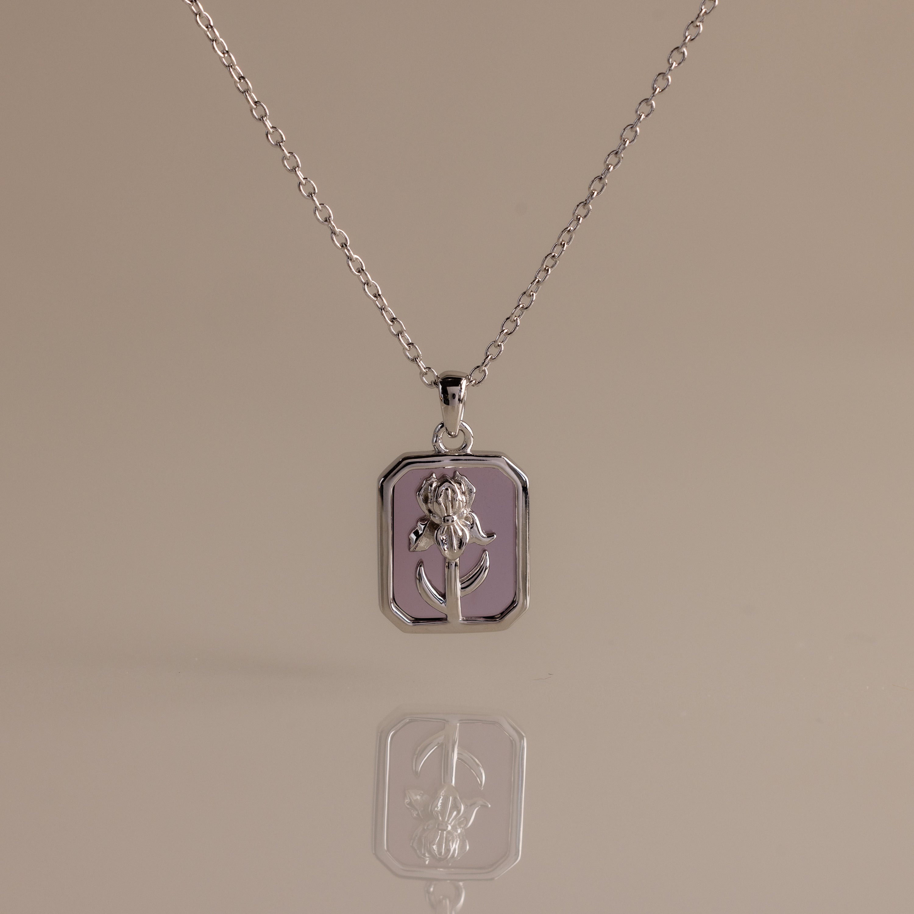 The Amethyst Birth Flower Charm Necklace features a silver rectangular pendant with a raised floral design, making it a perfect delicate jewelry piece. Displayed on a neutral background for an elegant presentation.