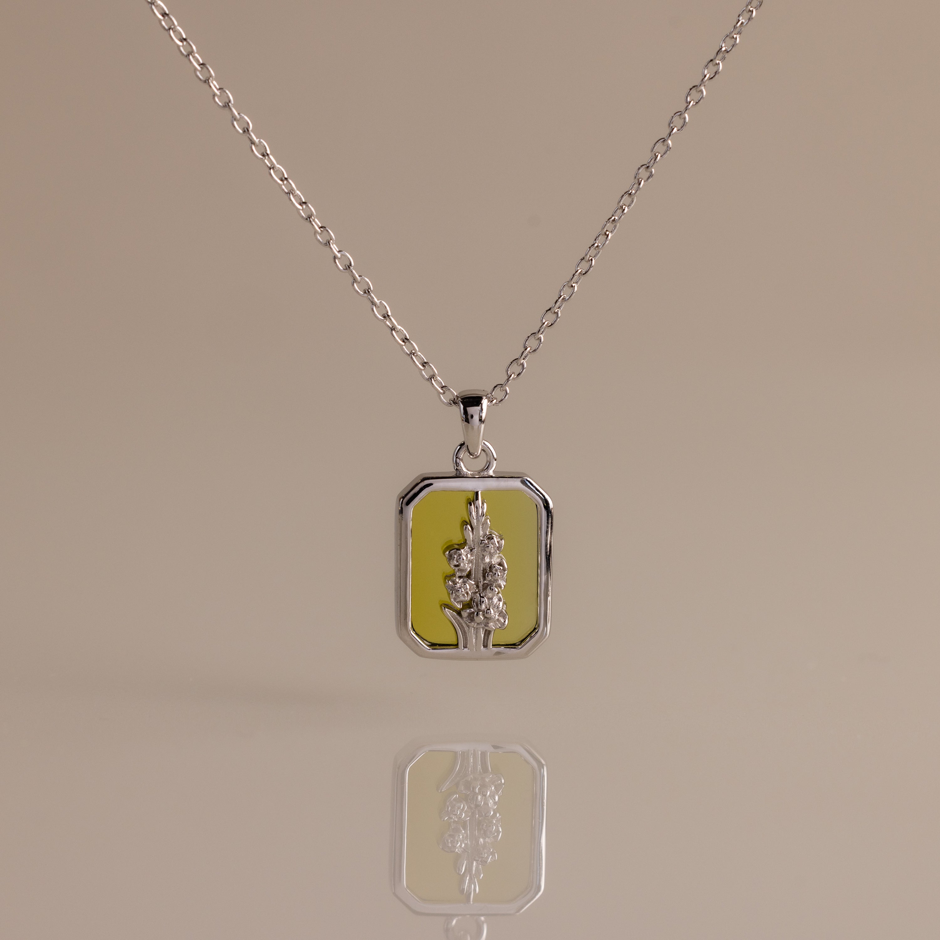 The Peridot Birth Flower Charm Necklace features a rectangular floral pendant on a yellow background, making it an ideal gift for nature lovers and flower jewelry enthusiasts. Displayed on a plain surface.
