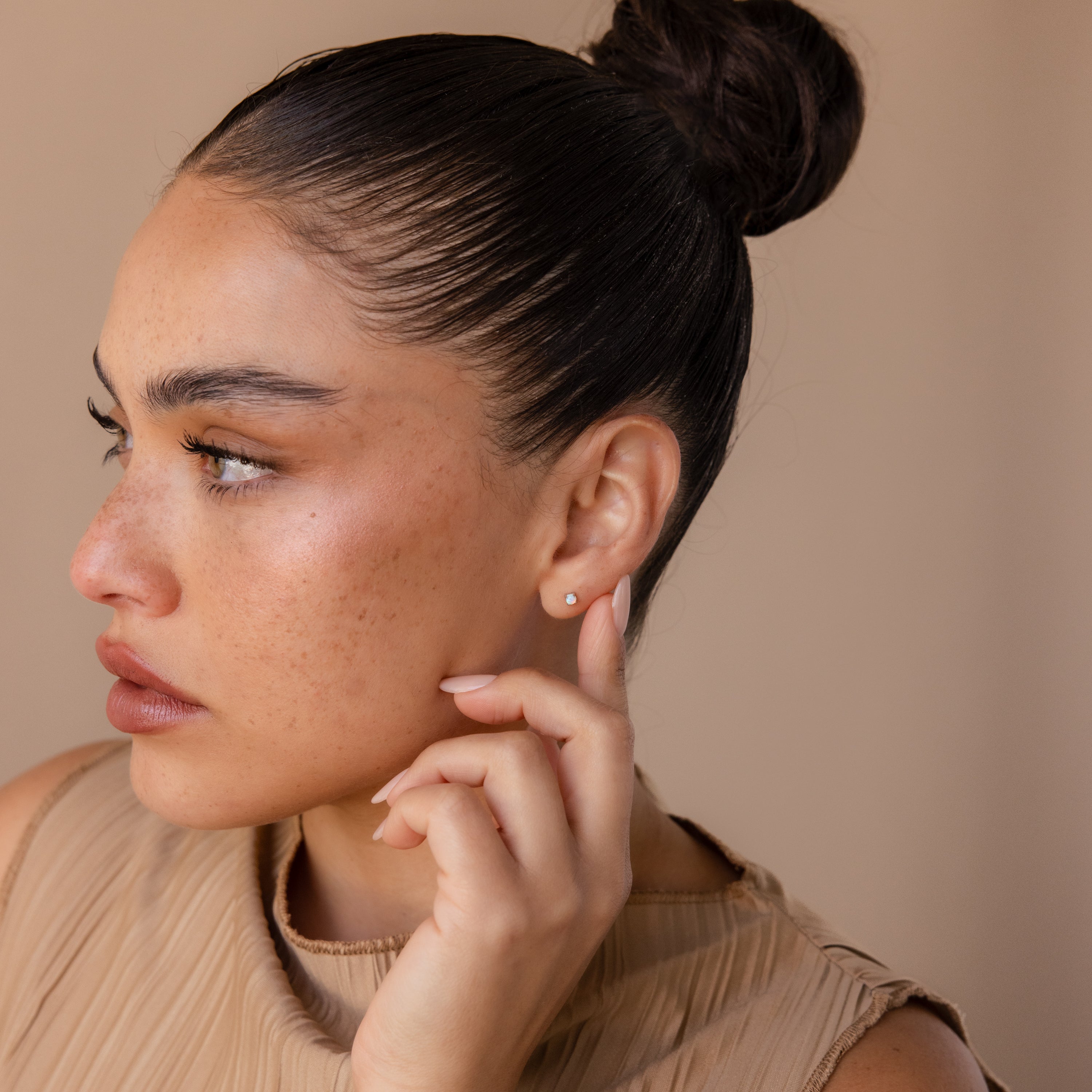 Woman with slicked-back hair and neutral makeup, touching her ear adorned with comfortable Opal Flat Back Studs in Sterling Silver, wearing a beige top and looking left.