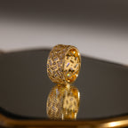 The Mosaic Diamond Baguette Ring features a bold, geometric cutout design in gold, beautifully displayed as its wide band elegantly reflects on a shiny surface.