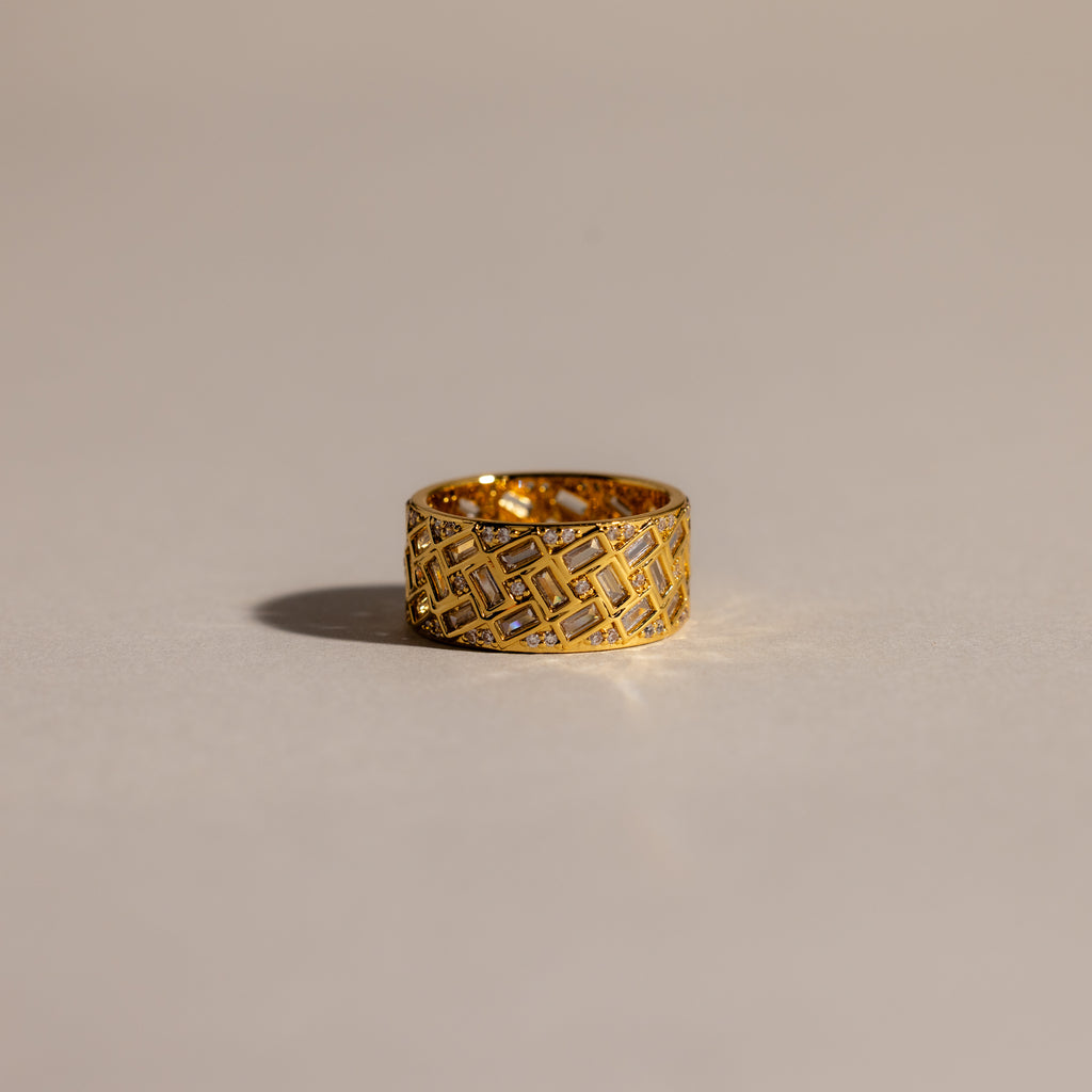 The Mosaic Diamond Baguette Ring, featuring a bold gold band with a geometric engraved pattern, is displayed on a softly lit, neutral surface.