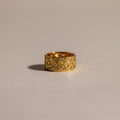 The Mosaic Diamond Baguette Ring, featuring a bold gold band with a geometric engraved pattern, is displayed on a softly lit, neutral surface.