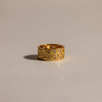 The Mosaic Diamond Baguette Ring, featuring a bold gold band with a geometric engraved pattern, is displayed on a softly lit, neutral surface.