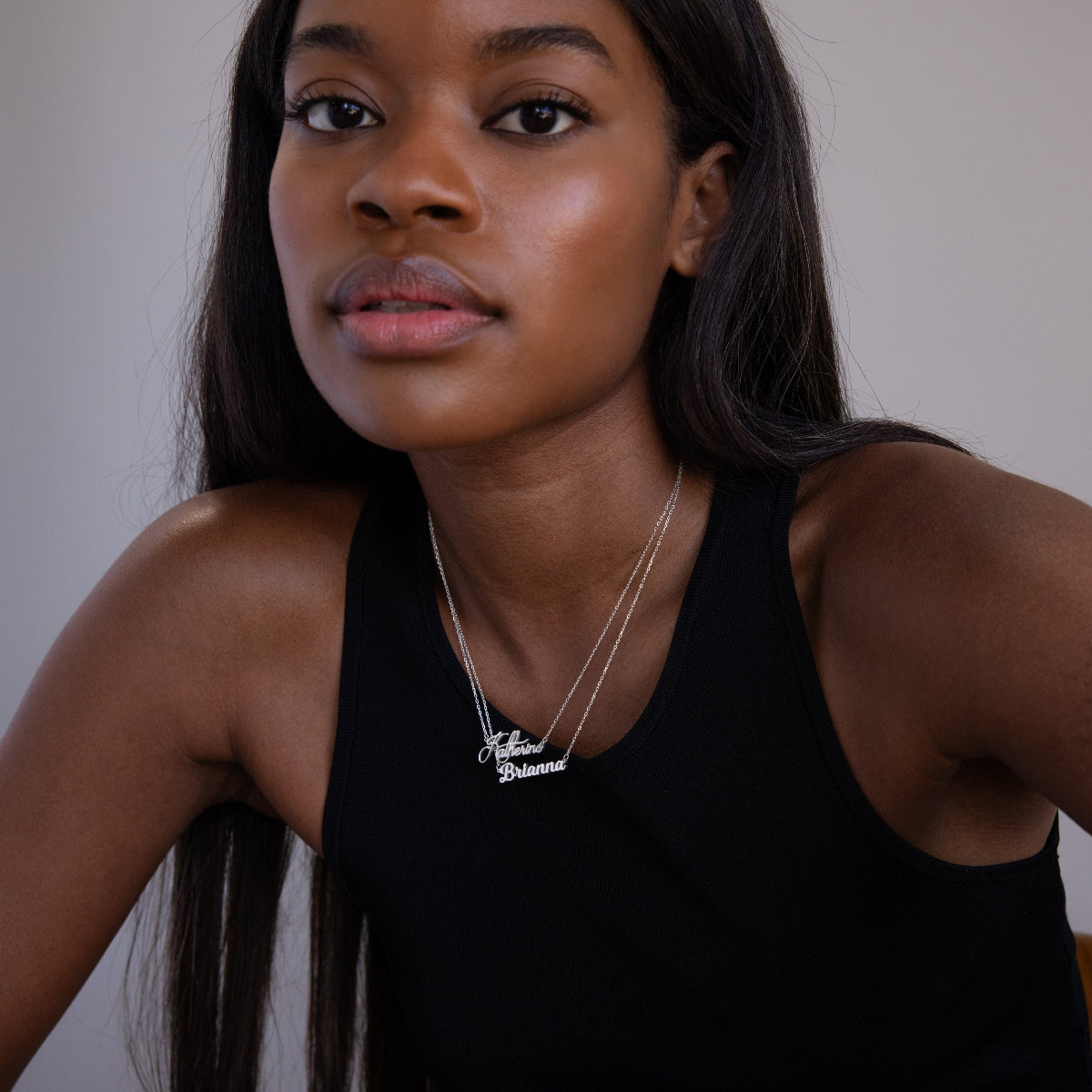 Woman with long dark hair wearing a black tank top and layered silver necklaces, including the chic Mellow Name Necklace in Sterling Silver, looks at the camera.