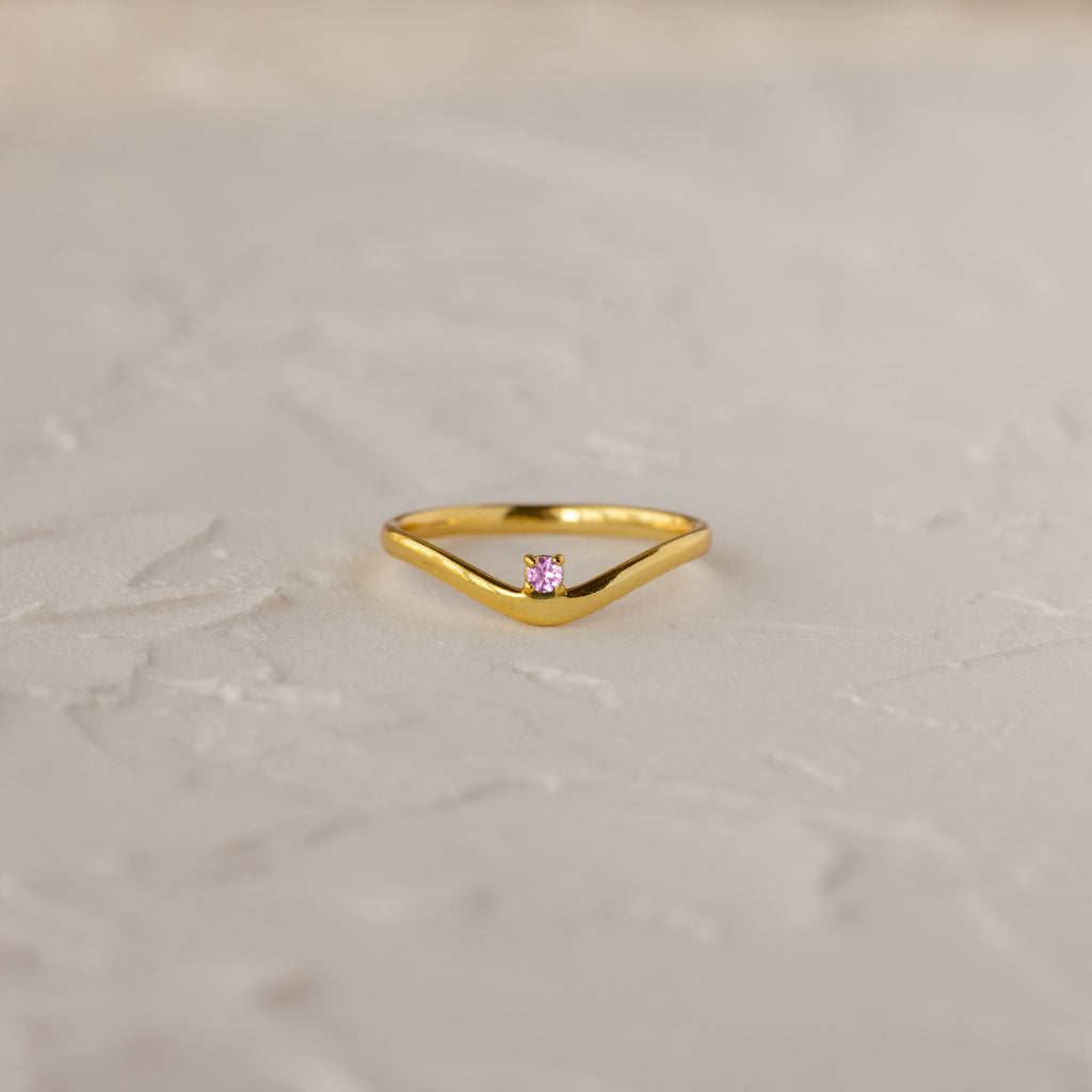 The Pink Tourmaline Drop Curve Ring is a delicate gold stacking ring featuring a small pink gemstone, shown on a textured white surface.