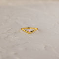 The Pink Tourmaline Drop Curve Ring is a delicate gold stacking ring featuring a small pink gemstone, shown on a textured white surface.
