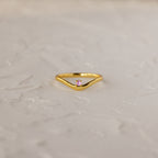 The Pink Tourmaline Drop Curve Ring is a delicate gold stacking ring featuring a small pink gemstone, shown on a textured white surface.