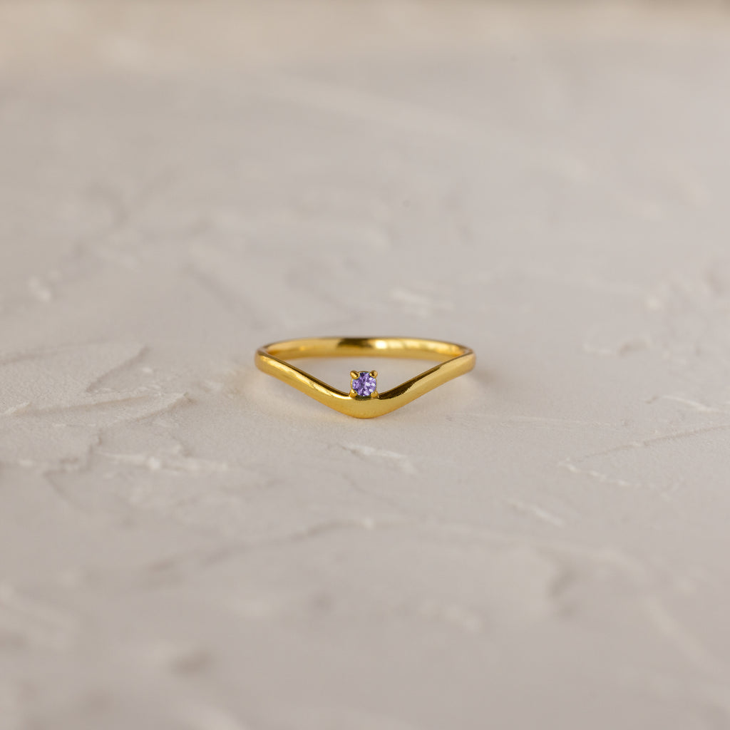 The Alexandrite Drop Curve Ring, featuring a delicate gold band with an alexandrite stone at the center, is displayed on a textured white surface.