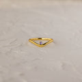The Alexandrite Drop Curve Ring, featuring a delicate gold band with an alexandrite stone at the center, is displayed on a textured white surface.