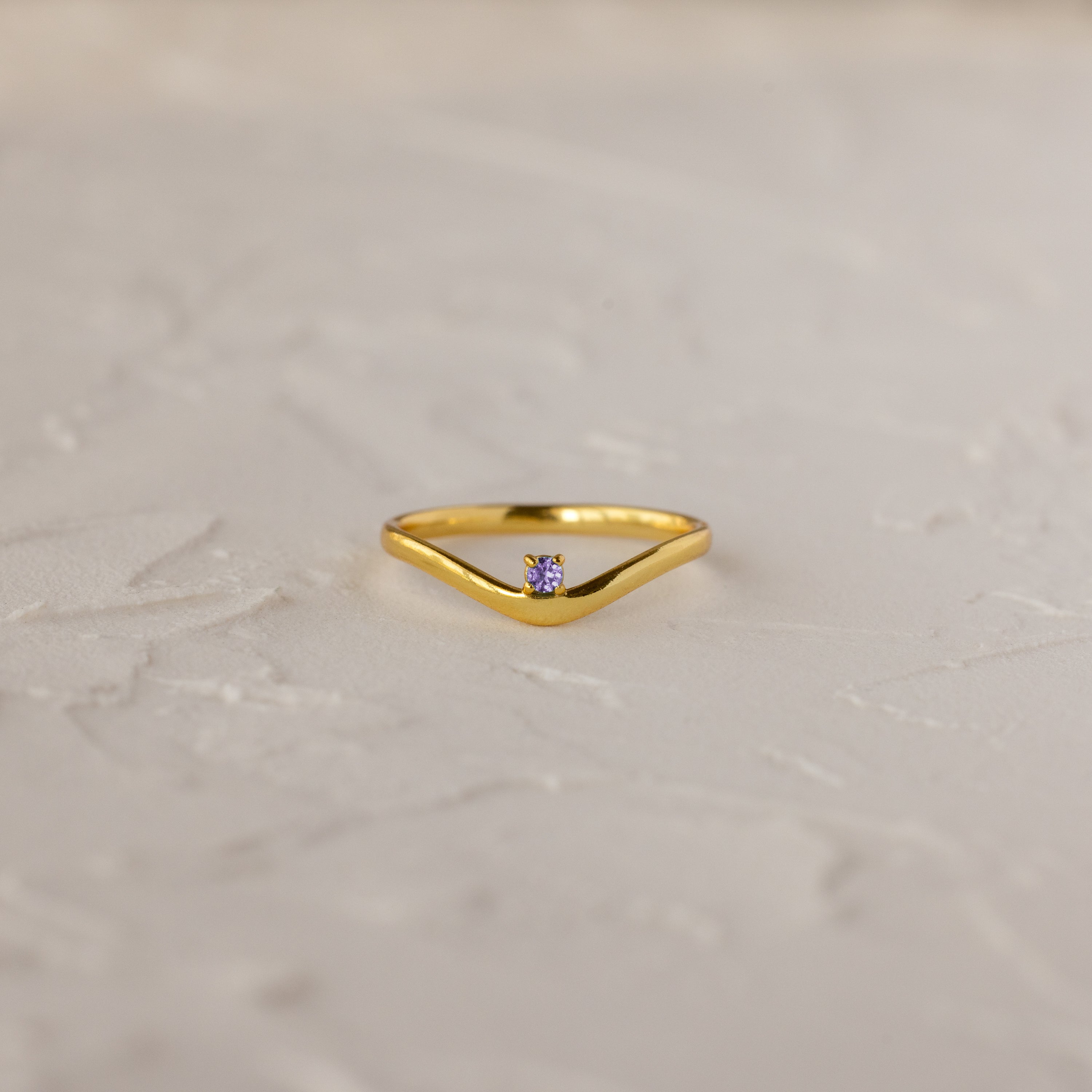 The Alexandrite Drop Curve Ring, featuring a delicate gold band with an alexandrite stone at the center, is displayed on a textured white surface.