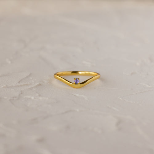 The Alexandrite Drop Curve Ring, featuring a delicate gold band with an alexandrite stone at the center, is displayed on a textured white surface.