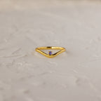 The Amethyst Drop Curve Ring features a delicate gold band with an amethyst accent, showcased on a textured white surface.