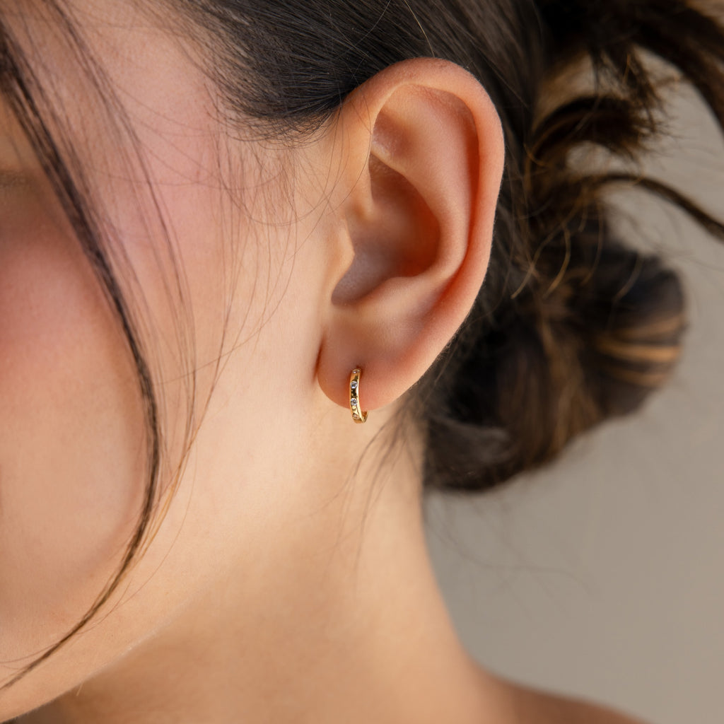 Close-up of a womans ear wearing Mini Diamond Sprinkle Huggies in 18K Gold, hair tied back with a loose strand softly framing her face.