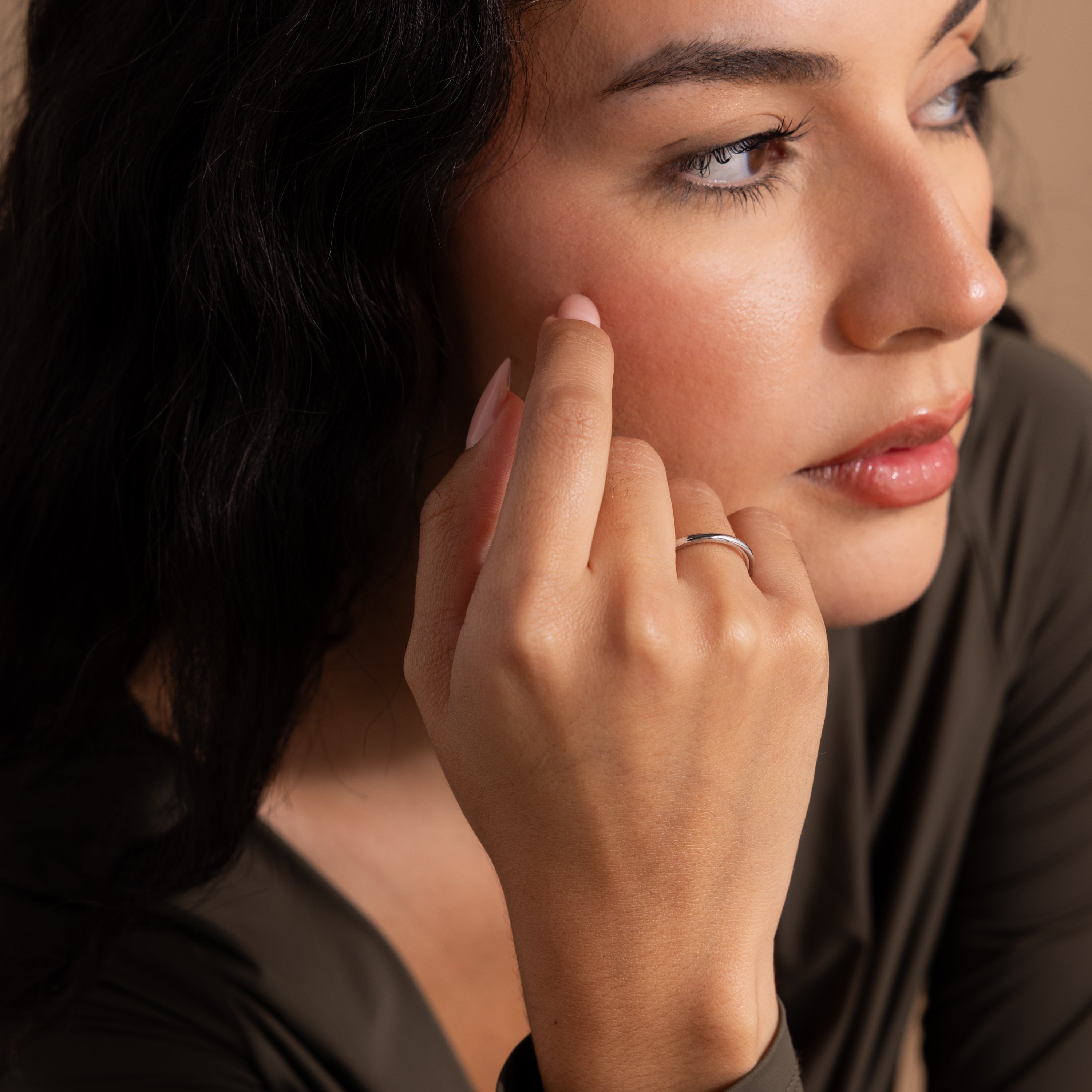 A woman with long dark hair, wearing a dark top and the Minimal Stacking Ring, touches her cheek while looking to the side.