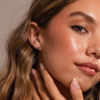 Woman with wavy brown hair wears Shark Tooth Flatback Studs in Silver and touches her face, showcasing natural makeup—a perfect look for a subtle beach-inspired accessory.