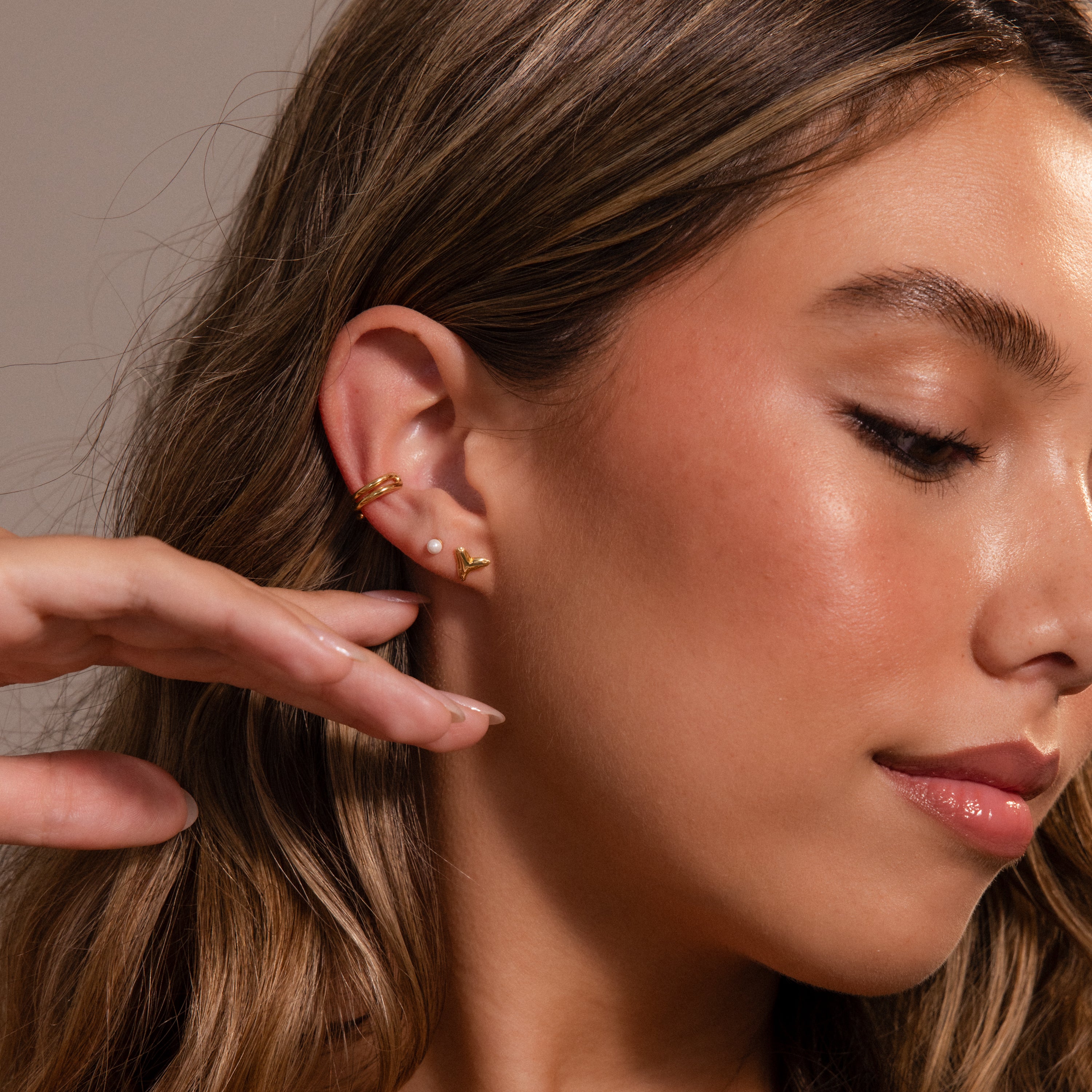 Woman with wavy brown hair wears Shark Tooth Flatback Studs in 18K Gold, a pearl earring, and a chic beach accessory, touching her face with manicured hand.