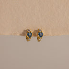 Aquamarine Heart Hoops: Gold earrings with blue heart-shaped stones and small clear gems, displayed on a textured beige surface—perfect as elegant birthstone earrings.