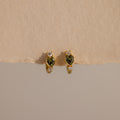 Peridot Heart Hoops featuring heart-shaped green stones and small clear accents, displayed on a beige surface—ideal as birthstone earrings or stylish gemstone huggies for any occasion.