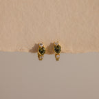 Peridot Heart Hoops featuring heart-shaped green stones and small clear accents, displayed on a beige surface—ideal as birthstone earrings or stylish gemstone huggies for any occasion.