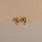Topaz Heart Hoops are birthstone earrings featuring heart-shaped topaz gems with small clear stones, shown on a textured beige background.